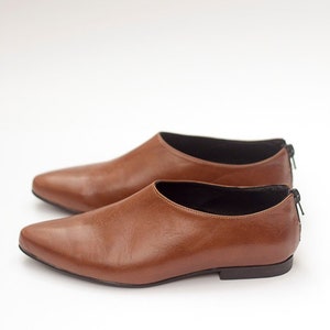 May include: A pair of brown leather slip-on shoes with pointed toes. The shoes have a black lining and a black zipper on the back.