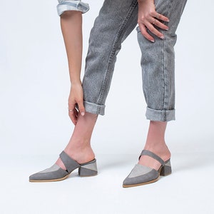 May include: A woman wearing a striped blue and white shirt, gray jeans, and gray heeled mules with a strap across the foot.