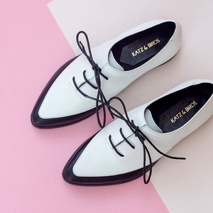 May include: Pair of white leather lace-up shoes with black trim and black laces. The shoes have a pointed toe and a low heel. The brand name "KATZ & BIRDS" is printed on the inside of each shoe.