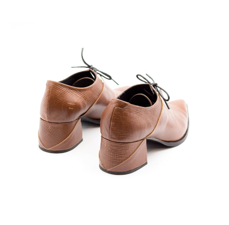 Op de afbeelding: Een paar bruine leren hakschoenen met zwarte veters. De schoenen hebben een uniek blokhakontwerp.