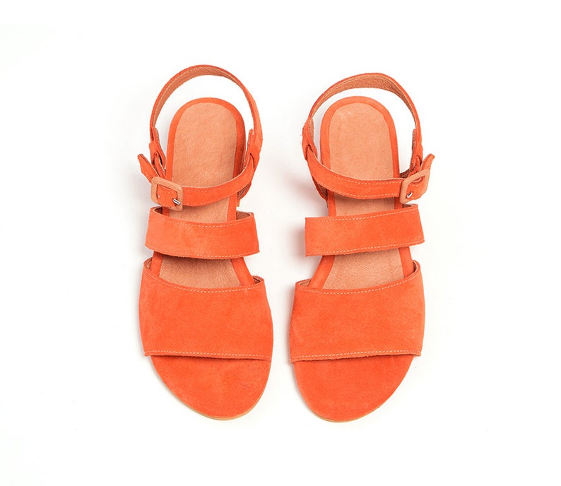 K&ouml;nnte beinhalten: Ein Paar orangefarbene Wildledersandalen mit drei Riemen und Schnallen. Die Sandalen haben eine flache Sohle und eine offene Zehenpartie.