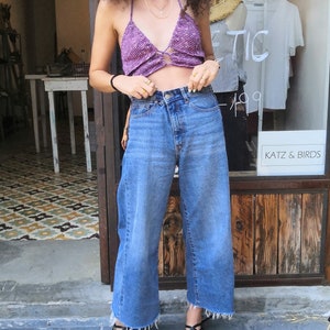 May include: A woman with curly brown hair is wearing a purple crochet top, blue denim wide-leg pants, and black sandals. She is standing in front of a store with a sign that says "Summer Up Tic 149".