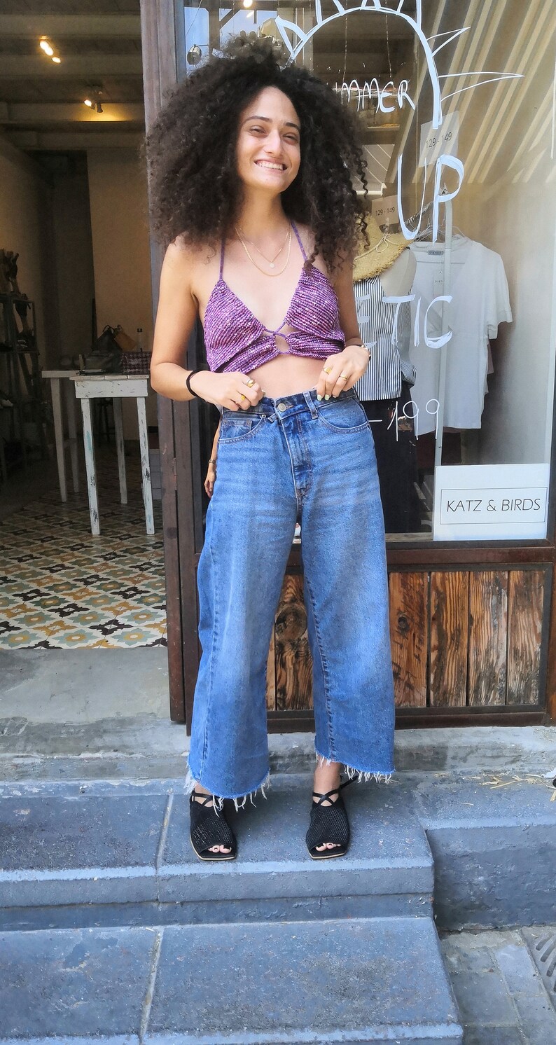 May include: A woman with curly brown hair is wearing a purple crochet top, blue denim wide-leg pants, and black sandals. She is standing in front of a store with a sign that says "Summer Up Tic 149".