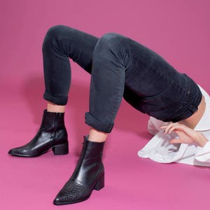 以下が含まれることがあります： 黒革のポインテッドトゥのアンクルブーツとブロックヒール。ブーツは濃い色のデニムジーンズを履いた人が履いています。