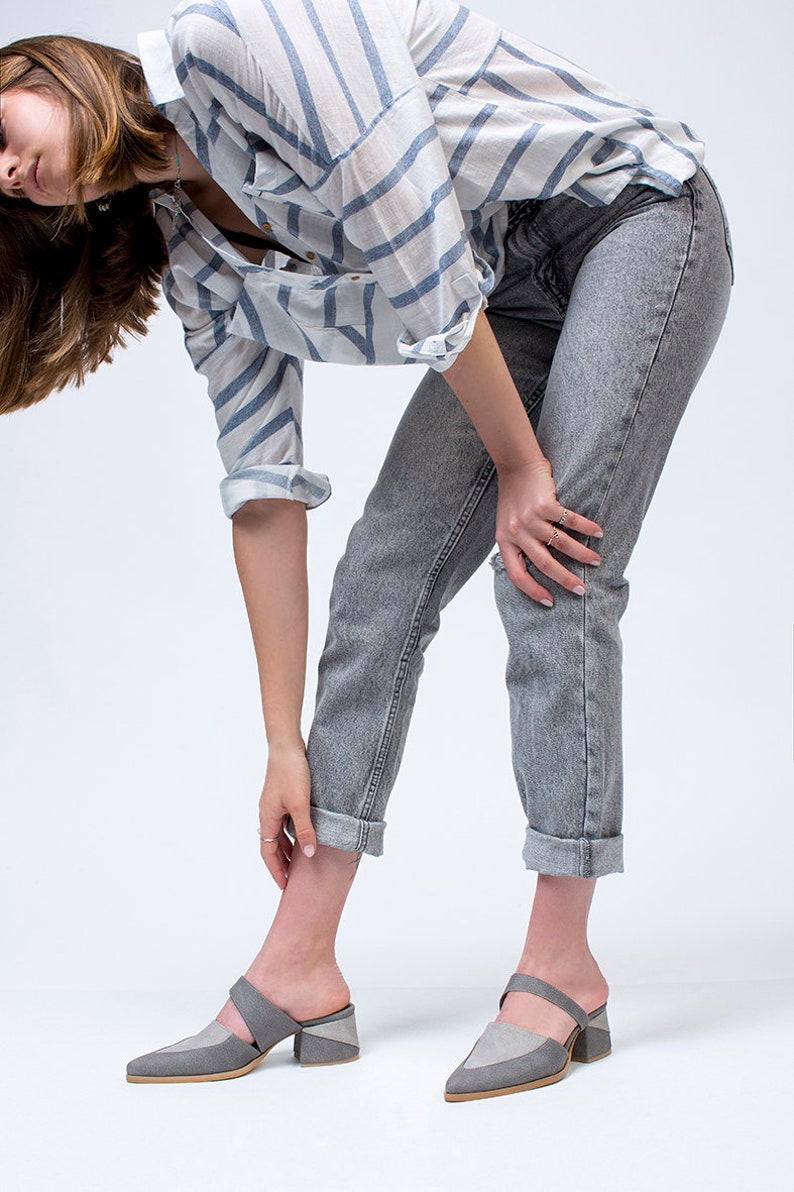 May include: A woman wearing a striped blue and white shirt, gray jeans, and gray heeled mules with a strap across the foot.