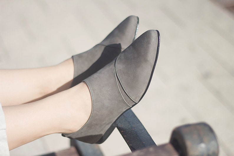 May include: A pair of gray suede shoes with pointed toes. The shoes are worn by a person with their legs crossed.
