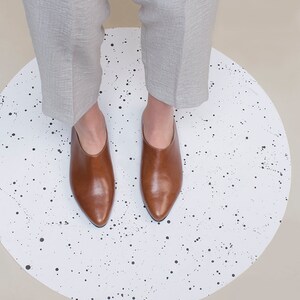 May include: A pair of brown leather heeled mules with pointed toes on a white background with black splatter paint.