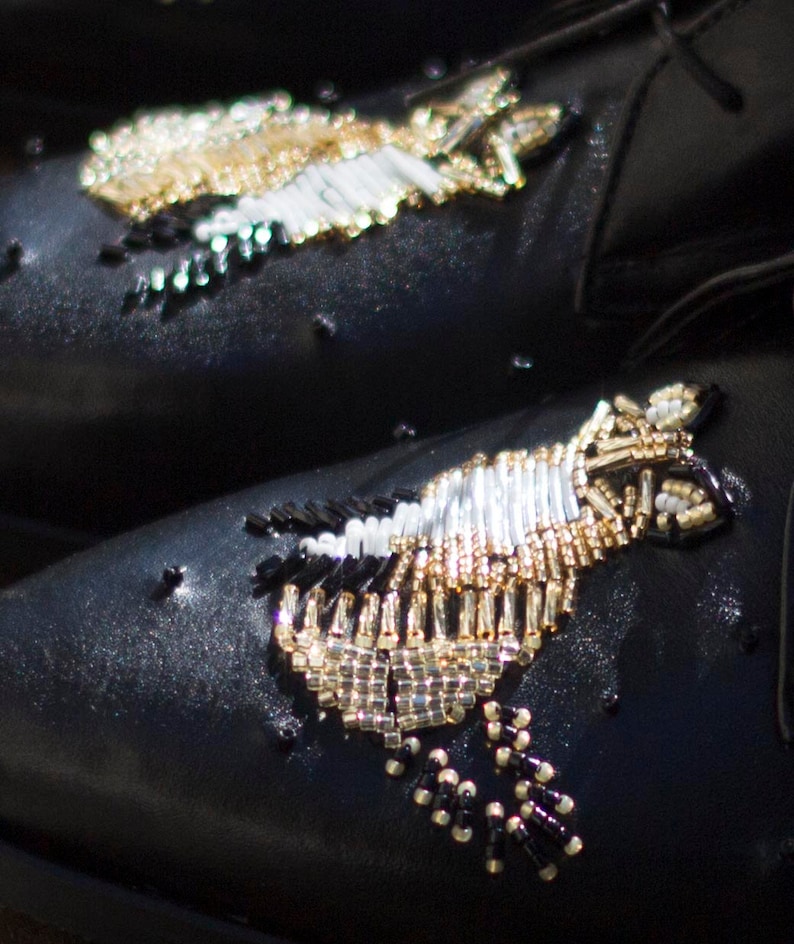 Puede incluir: Un par de zapatos de cuero negro con intrincados adornos de cuentas. Las cuentas est&aacute;n dispuestas en un patr&oacute;n que se asemeja a un p&aacute;jaro o un pez. Los zapatos tienen una punta puntiaguda y un tac&oacute;n bajo.