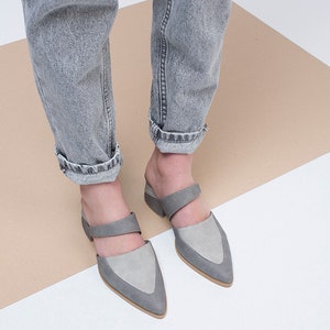 May include: A pair of gray suede heeled mules with a pointed toe and a strap across the top of the foot. The shoes are worn by a person wearing gray jeans.