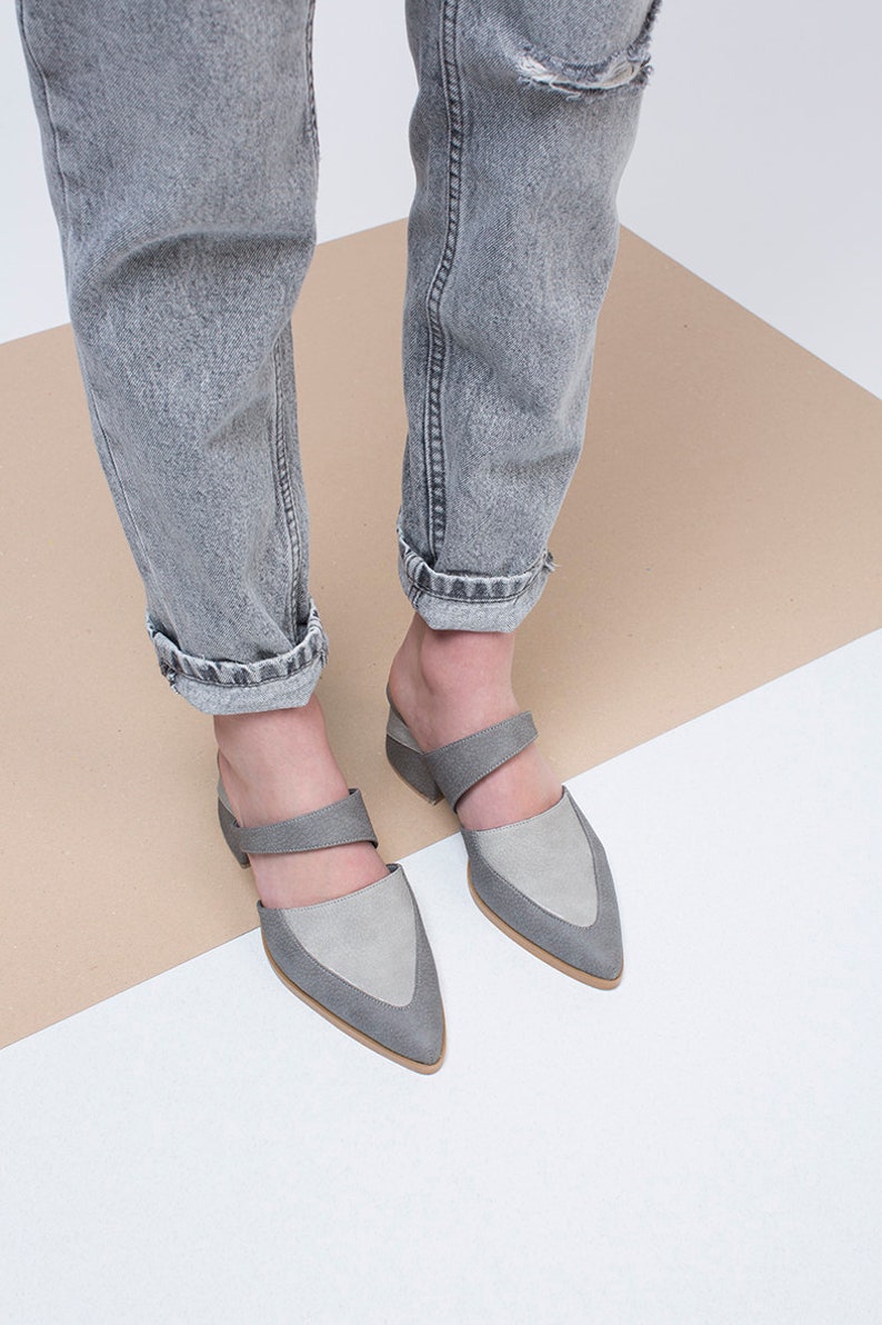 May include: A pair of gray suede heeled mules with a pointed toe and a strap across the top of the foot. The shoes are worn by a person wearing gray jeans.