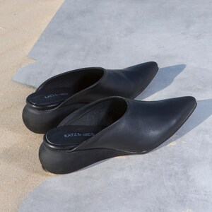 May include: A pair of black leather heeled mules with pointed toes. The shoes have a thick rubber sole and the brand name "KATZ & BIRDS" is printed on the insole.