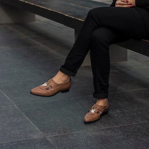Pode incluir: Um par de sapatos de couro marrom com bordados florais. Os sapatos t&ecirc;m bico fino e salto baixo.