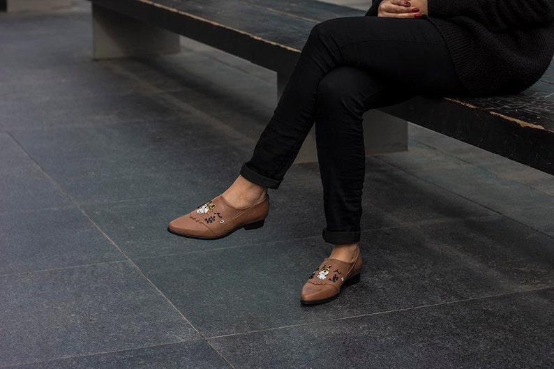 Pode incluir: Um par de sapatos de couro marrom com bordados florais. Os sapatos t&ecirc;m bico fino e salto baixo.
