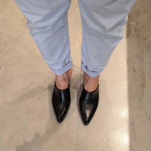 May include: A pair of black leather mules with pointed toes. The shoes are worn with light blue cuffed pants. The image is taken from a top-down perspective, showcasing the footwear and the lower portion of the legs.
