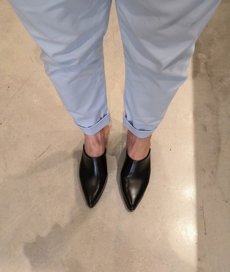 May include: A pair of black leather mules with pointed toes. The shoes are worn with light blue cuffed pants. The image is taken from a top-down perspective, showcasing the footwear and the lower portion of the legs.