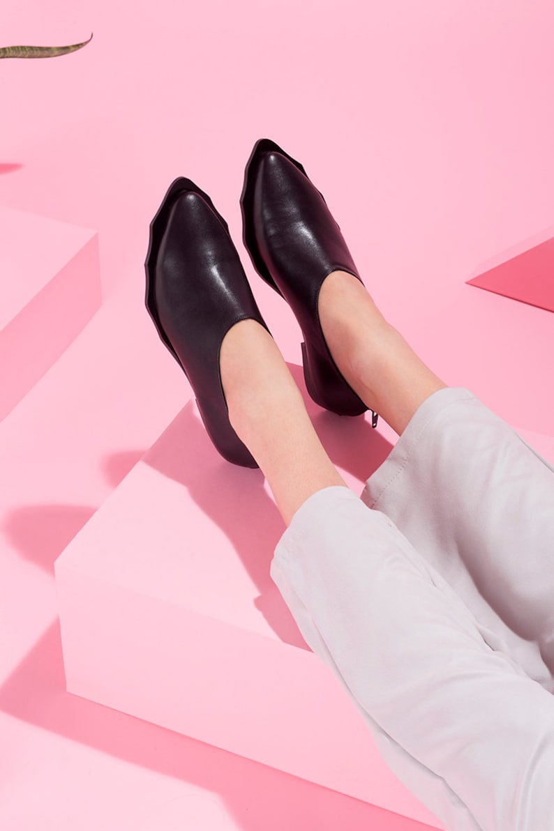 May include: A pair of black leather pointed-toe shoes with a unique scalloped edge design. The shoes are worn by a person with white pants on a pink background.