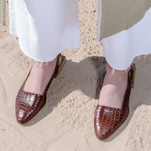 Op de afbeelding: Een paar bruine leren sandalen met hak en een krokodillenpatroon. De sandalen hebben een slingback-bandje en worden gedragen aan de voeten.