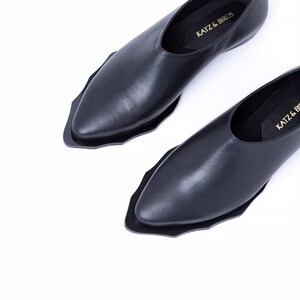 May include: A pair of black leather slip-on shoes with a pointed toe and a unique scalloped edge. The shoes have a simple design and feature the text "Katz & Birds" on the insole.