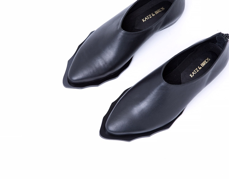 May include: A pair of black leather slip-on shoes with a pointed toe and a unique scalloped edge. The shoes have a simple design and feature the text "Katz & Birds" on the insole.