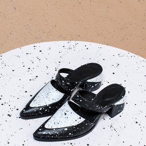 May include: Two black and white heeled mules with a splatter paint design. The shoes have a pointed toe and a thick heel.