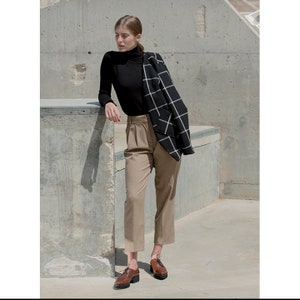 K&ouml;nnte beinhalten: Eine Frau tr&auml;gt einen schwarzen Rollkragenpullover, eine beige Hose und braune Lederschn&uuml;rschuhe. Sie tr&auml;gt einen schwarz-wei&szlig; karierten Blazer &uuml;ber ihrem Pullover.