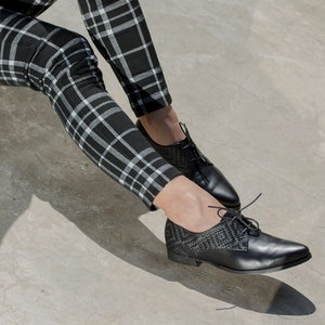 Op de afbeelding: Zwarte leren veterschoenen met een geweven detail op de neus en hiel. De schoenen worden gedragen met een zwart-witte geruite broek.