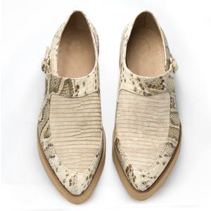 Puede incluir: Un par de zapatos de cuero beige con punta puntiaguda y estampado de piel de serpiente. Los zapatos tienen un cierre de hebilla y una suela gruesa.