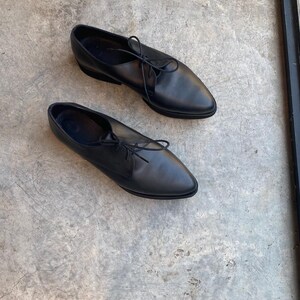 May include: A pair of black leather lace-up shoes with pointed toes. The shoes are on a grey concrete surface.