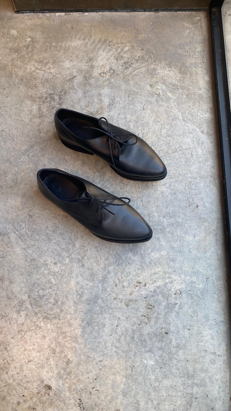 May include: A pair of black leather lace-up shoes with pointed toes. The shoes are on a grey concrete surface.