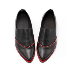 May include: A pair of black leather shoes with a pointed toe and a red trim around the edge. The shoes have a zipper on the back.