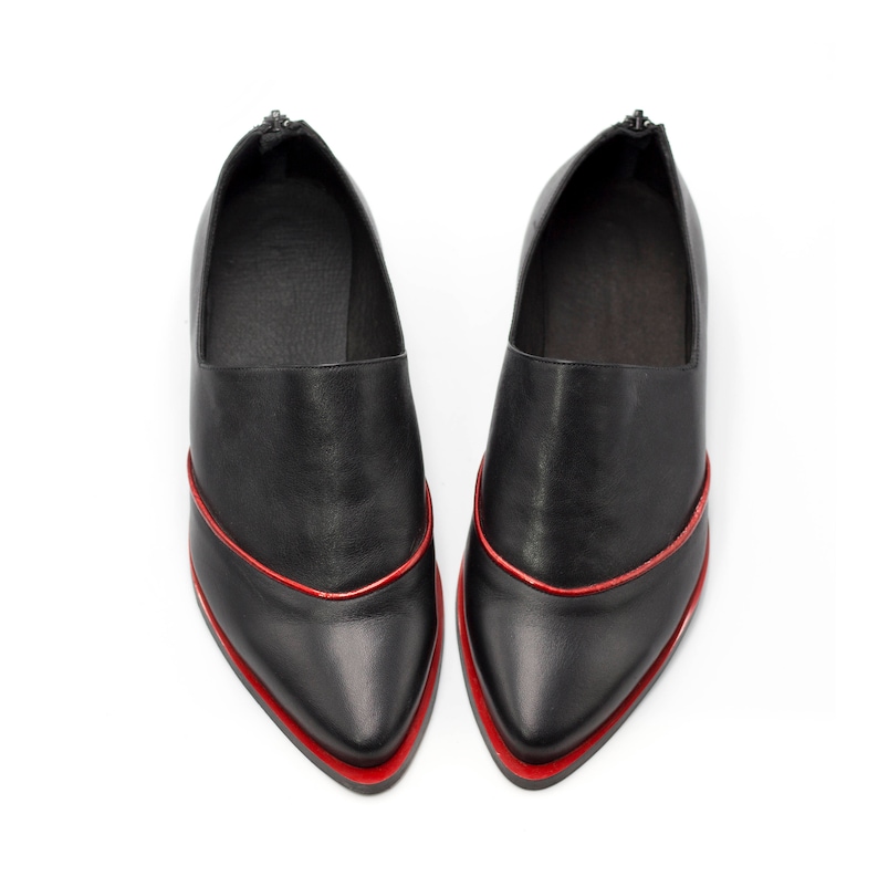 May include: A pair of black leather shoes with a pointed toe and a red trim around the edge. The shoes have a zipper on the back.