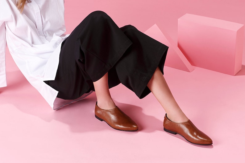 May include: A pair of brown leather slip-on shoes with pointed toes. The shoes are worn by a person sitting on a pink surface.