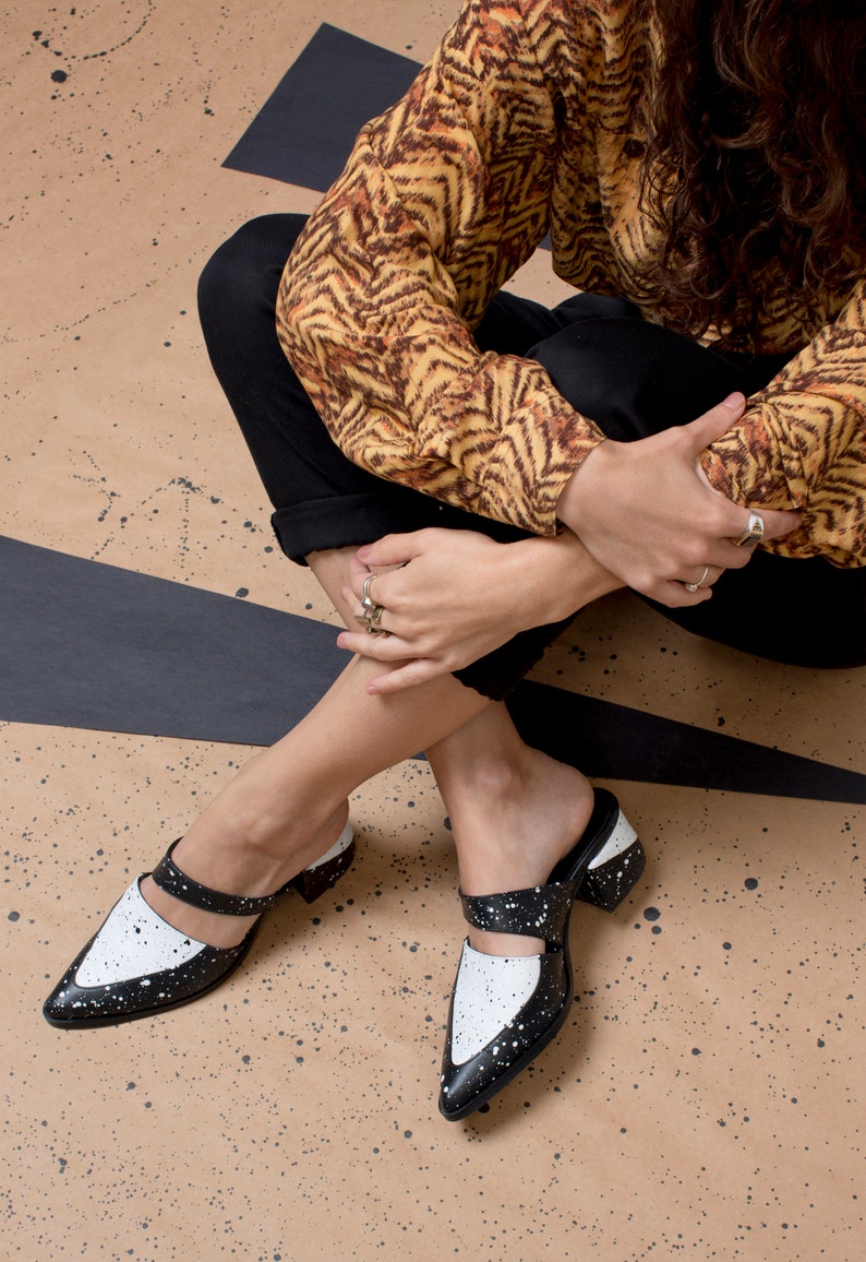May include: A pair of black and white heeled mules with a splatter paint design. The shoes have a pointed toe and a strap across the top of the foot.