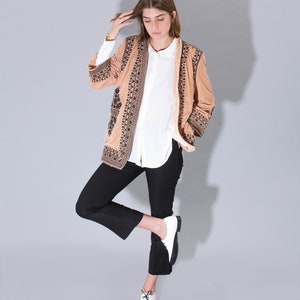 May include: A woman wearing a brown embroidered jacket with black trim, a white button-down shirt, black pants, and white and black shoes.