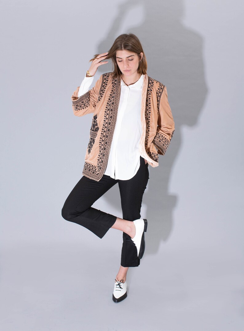 May include: A woman wearing a brown embroidered jacket with black trim, a white button-down shirt, black pants, and white and black shoes.