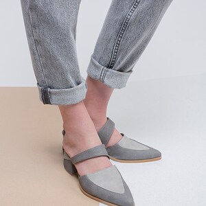 May include: A pair of gray suede heeled shoes with a pointed toe and a strap across the foot. The shoes are worn by a person wearing gray jeans.