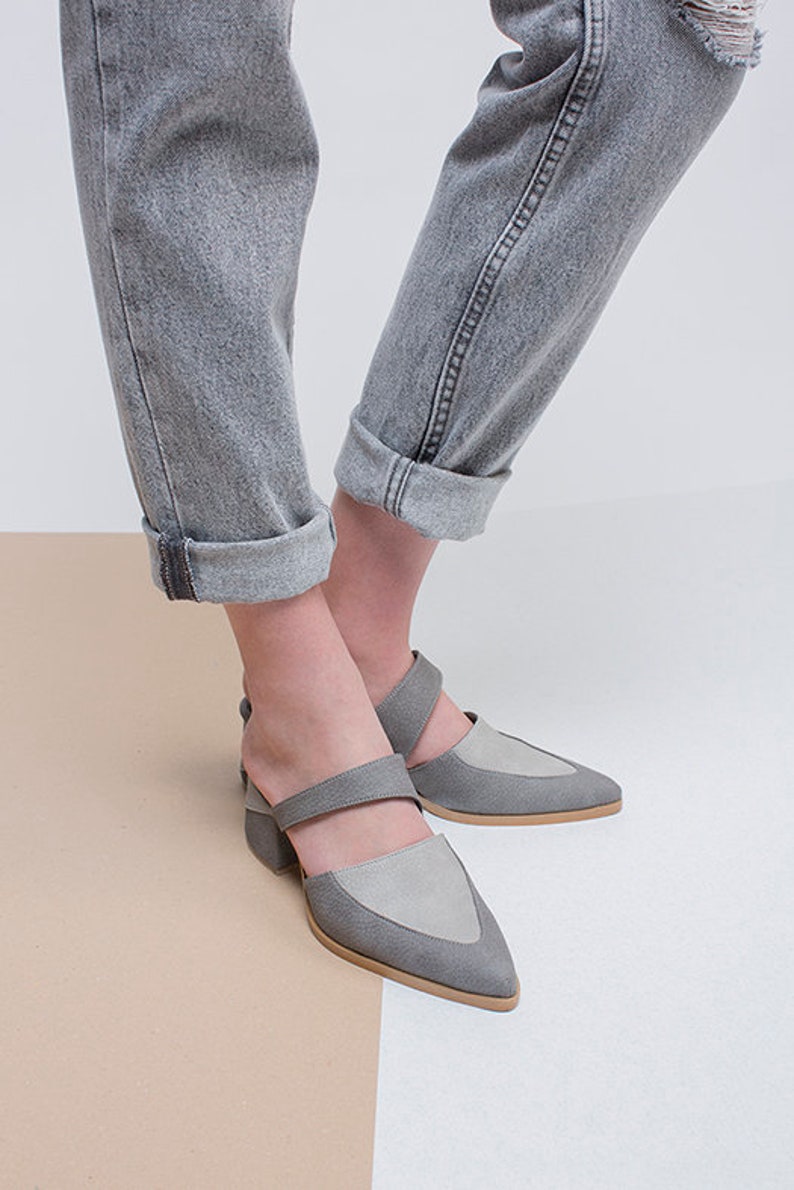May include: A pair of gray suede heeled shoes with a pointed toe and a strap across the foot. The shoes are worn by a person wearing gray jeans.