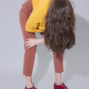 May include: A person wearing a mustard yellow sweater, rust-colored pants, and shiny red leather shoes with black laces. The person is bent over, with long brown hair covering their face.