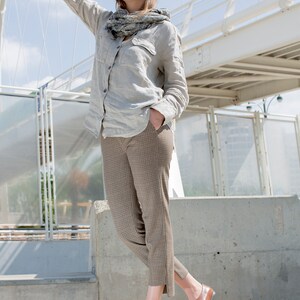 May include: A woman wearing a light gray linen shirt with a gray and white scarf, brown plaid pants, and brown leather shoes. She is standing on a concrete surface with a white bridge in the background.