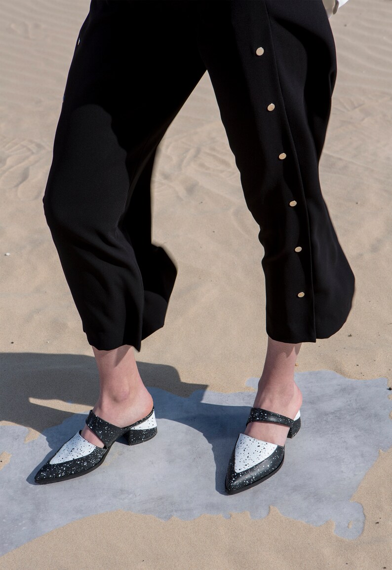 May include: A pair of black and white heeled mules with a pointed toe. The shoes have a white base with black speckles and a black strap across the top.