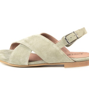 K&ouml;nnte beinhalten: Ein Paar hellgr&uuml;ne Wildledersandalen mit einem Kreuzriemen-Design und einem Schnallenverschluss am Kn&ouml;chelriemen. Die Sandalen haben eine flache Sohle und ein braunes Lederfu&szlig;bett.