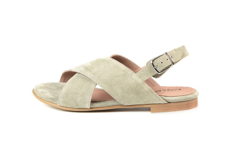 K&ouml;nnte beinhalten: Ein Paar hellgr&uuml;ne Wildledersandalen mit einem Kreuzriemen-Design und einem Schnallenverschluss am Kn&ouml;chelriemen. Die Sandalen haben eine flache Sohle und ein braunes Lederfu&szlig;bett.