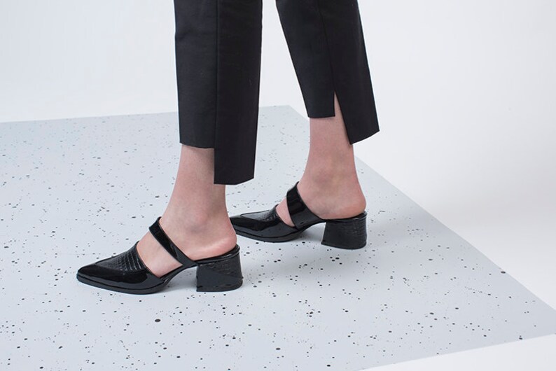 May include: Black heeled mules with a pointed toe and two straps. The shoes have a textured, crocodile-embossed design.
