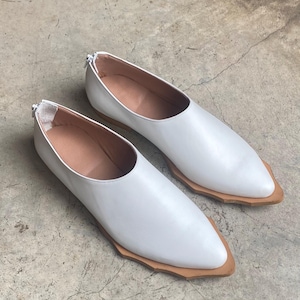 May include: A pair of white slip-on shoes with a pointed toe and a tan, jagged-edged sole. The shoes feature a smooth white upper, a tan interior lining, and a small silver zipper closure located at the back of the heel collar for easy access.