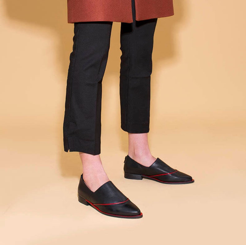 Puede incluir: Zapatos de vestir negros de cuero con un detalle de costura roja. Los zapatos tienen una punta puntiaguda y un tac&oacute;n bajo.
