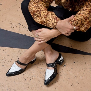 May include: Black and white leather heeled mules with a pointed toe and a strap across the foot. The shoes have a white splatter paint design.