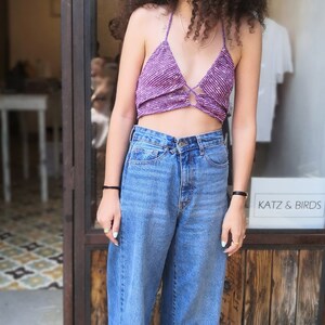 May include: A woman wearing a purple crochet halter top with a tie detail, blue denim wide-leg jeans, and black sandals. She is standing in front of a store window with the text "Katz & Birds" visible.