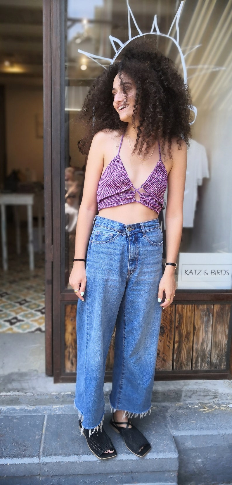 May include: A woman wearing a purple crochet halter top with a tie detail, blue denim wide-leg jeans, and black sandals. She is standing in front of a store window with the text "Katz & Birds" visible.
