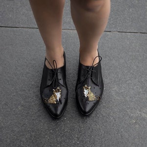 Puede incluir: Zapatos negros de cuero con cordones, punta puntiaguda y adornos de cuentas doradas y blancas en la parte delantera.