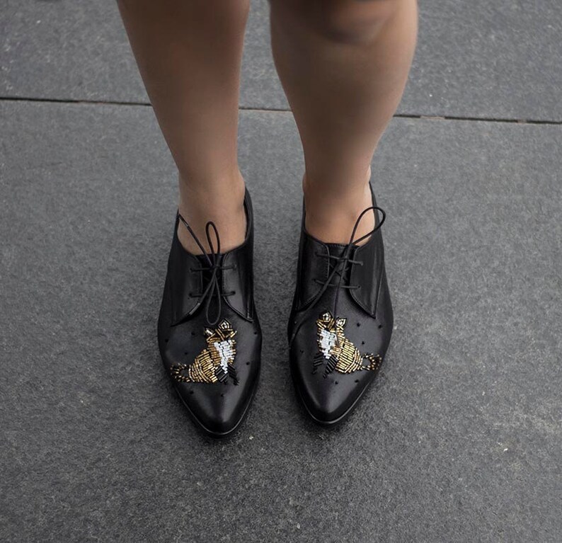 Puede incluir: Zapatos negros de cuero con cordones, punta puntiaguda y adornos de cuentas doradas y blancas en la parte delantera.